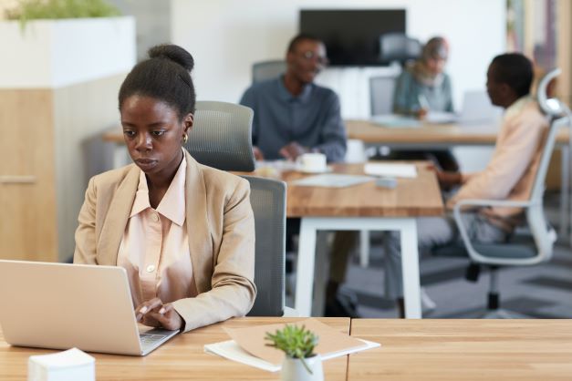 African American woman working on a laptop in an office with coworkers blurred behind her.